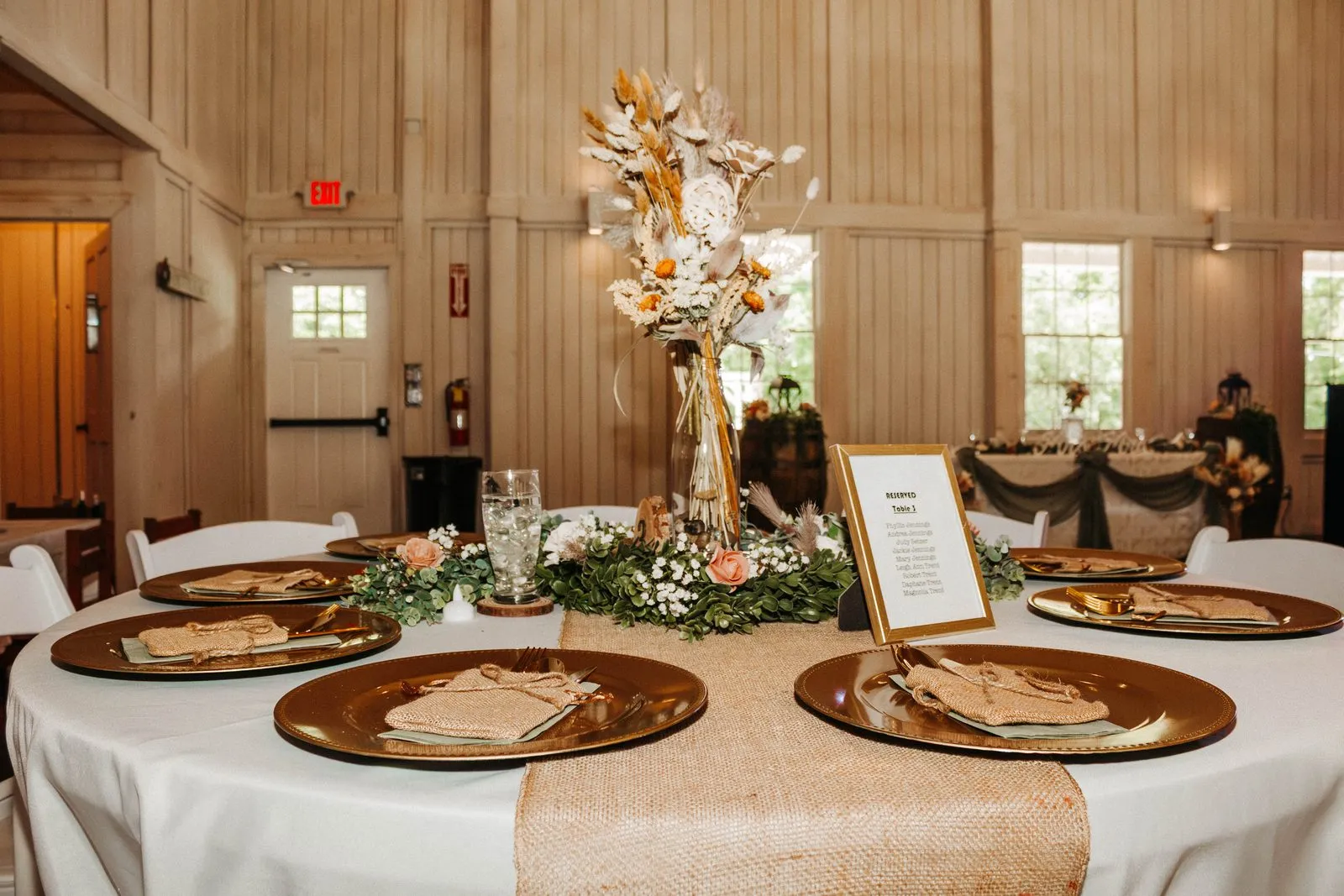 Round reception table with greenery, gold chargers, and a tall floral centerpiece at The Heritage Barn